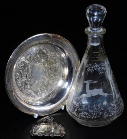 A plated dish, of circular form, 14cm diameter, a moulded glass decanter with etch style deer and tree emblem and a silver gin label. (3)