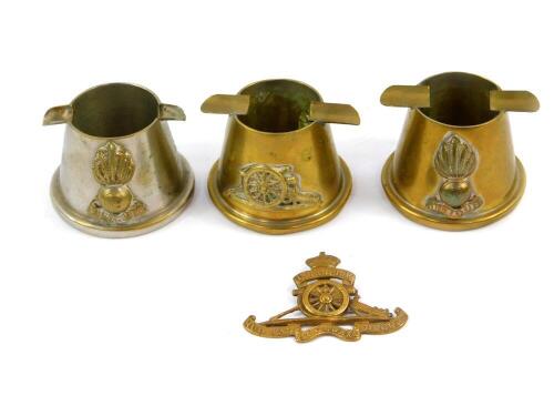 Three Royal Artillery trench art metal ashtrays, together with a Royal Artillery cap badge. (4)