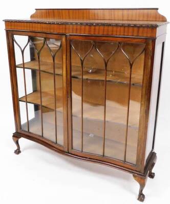 A 20thC mahogany serpentine display cabinet, the carved top raised above a pair of astragal glazed doors, on compressed ball and claw feet, 132cm high, 120cm wide, 33cm deep.