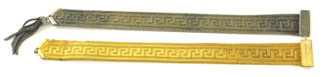 Two Eastern style dress tabs, each with Aztec type design to centre, one gold coloured the other silver coloured, gold coloured lacking tassle. (2)