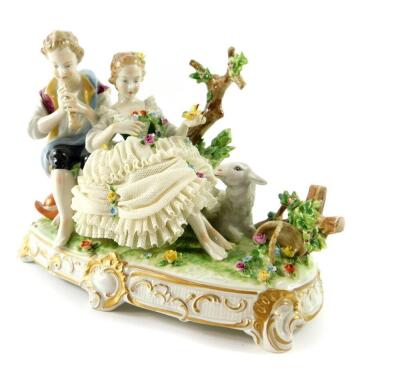 An Unterwiesbach 20thC porcelain figure group, modelled as a shepherd and shepherdess with lamb, both figures seated, the shepherd playing a woodwind instrument, the girl holding a wreath and canary, backed by bocage, raised on a naturalist oval rococo st