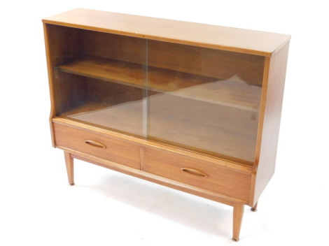 A mid century teak display cabinet, with two sliding glazed doors and single shelf, above a pair of frieze drawers, raised on turned legs, brass capped, 87cm high, 107cm wide, 30.5cm deep.