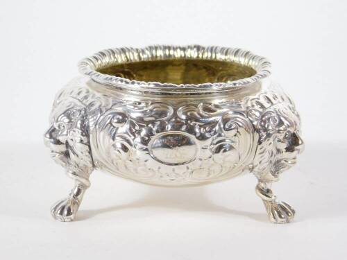 A George IV crested silver circular salt, with everted rim and swollen body with repousse scroll and flower head decoration, raised on three lions head and claw feet, London 1828, makers mark WB, 5.4oz.