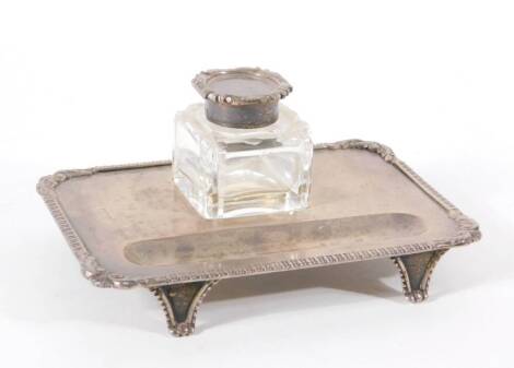 An Edwardian silver desk stand, of rectangular design with gadrooned border and foliate scroll corners, raised on four tapered feet, with silver capped cut glass ink receiver, London 1905, 168cm long, 6.3oz.