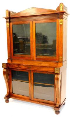 A Regency rosewood and brass cabinet, the top with an arched cornice flanked by lotus and acanthus motifs, above two glazed doors enclosing adjustable shelves, the base with a frieze drawer and two further glazed doors flanked by shaped supports on lobed