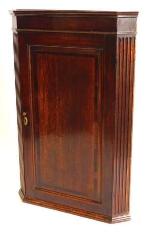 An early 19thC oak and mahogany cross banded hanging corner cabinet, with a single panelled door flanked by reeded canted sides, 106cm high, 69cm wide.