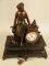 A French black marble and spelter mantel clock in the form of a lady seated beside birds