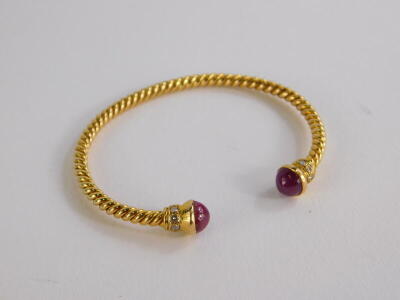 A torc bangle, the bangle ends set with cabochon cut ruby, and diamond surround, on a spiral turned bracelet, yellow metal, unmarked, 6cm wide, 16.3g all in. - 2