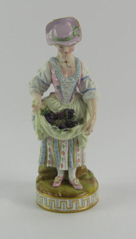 A Meissen porcelain figure of a lady, modelled standing in eighteenth century costume with grapes gathered in her apron, raised on a naturalistic circular base, 18cm high, (AF).