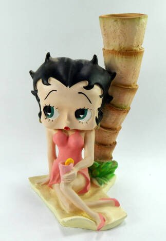 A plaster figure of Betty Boop, modelled seated on a bench with a drink, beside a palm tree, 49cm high.