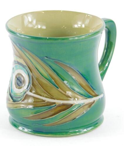 A Moorcroft pottery mug, decorated with a peacock feather, on a green ground, 8.5cm high.