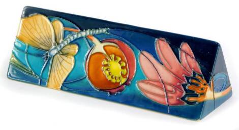 A Moorcroft pottery triangular plaque, decorated to one side with a dragonfly, fruits, flowers, etc., on a turquoise ground, 18.5cm wide.