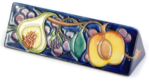 A Moorcroft pottery triangular plaque, decorated to one side with fruit, to include a peach, a fig and a raspberry, etc., on a blue ground, 18.5cm wide.