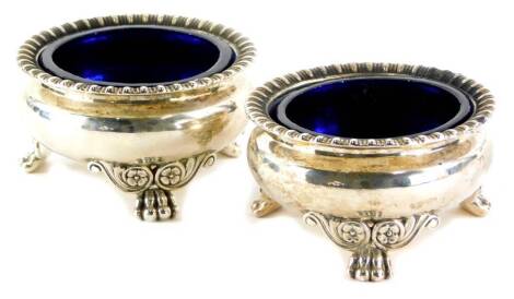 A pair of George III silver salts, each with a gadrooned border on paw feet, headed by scrolls, etc., blue glass liners, London 1817, weighable silver 9¼oz, 8.5cm diameter.