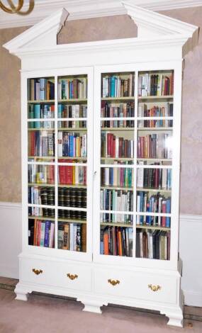 A white painted Neo-classical style bookcase, with a broken arch pediment, over a pair of glazed doors, enclosing five shelves, above two drawers, raised on ogee bracket feet, 280cm high, 173cm wide, 38cm deep.