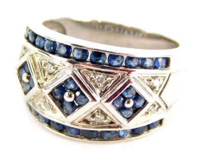 A sapphire and diamond set dress ring, with three central diamond motifs each set with round brilliant cut sapphires, forming a flower with pave set tiny diamonds, and an outer sapphire band border, white metal stamped 10k, ring size O, 5.8g all in, boxed