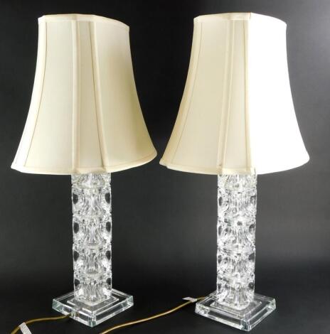 A pair of square section moulded glass table lamps with shades, the bases 48cm high to the top of the fitting.