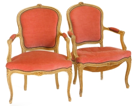 A pair of early 20thC French open armchairs, each with a padded back, armrest and seat, upholstered in pink fabric, the frame painted in cream, embellished in gilt, on cabriole legs.