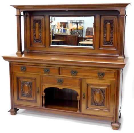 A late 19th/early 20thC walnut sideboard, the raised back with central bevelled mirror plate, flanked by carved panels on square section fluted supports, the base with two frieze drawers, above a central drawer and an arched recess, flanked by two panelle