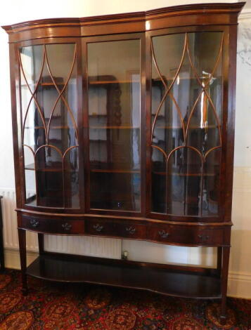 A late Victorian flamed mahogany display cabinet, with satinwood banded double bow front and serpentine cornice, two astragal glazed doors, flanking a central glazed panel, over three drawers with brass loupe handles, under shelf, square taper legs and sp