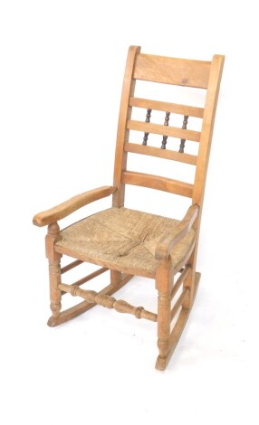 An early 20thC kitchen rocking chair, with horizontal back splats, some broken by inverted ball pieces, with shaped arms, rush seats and turned front legs, on a rocker base, 105cm high.