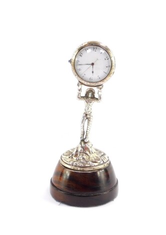 A Continental late 19thC novelty table clock, circular enamel dial bearing Arabic numerals, centre seconds, chased brass cased visible movement, the circular white metal casing foliate engraved, held aloft by a figure of Harlequin standing on a Rococo scr