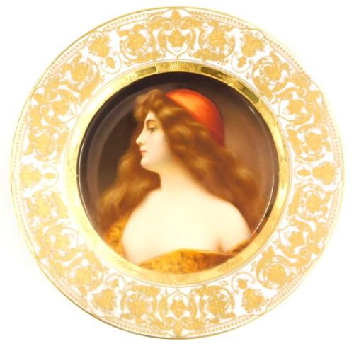 A 19thC Vienna porcelain cabinet plate, decorated with a portrait of a young lady with flowing brown hair and a red hat, titled Una Gitana, signed Wagner, within a pink and elaborate gilt border, 24.5cm diameter.