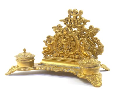 A Victorian rococo style brass desk stand, cast with cherubs, winged beasts and vase of flowers, having a letter rack pen tray and double ink wells, 30cm wide.