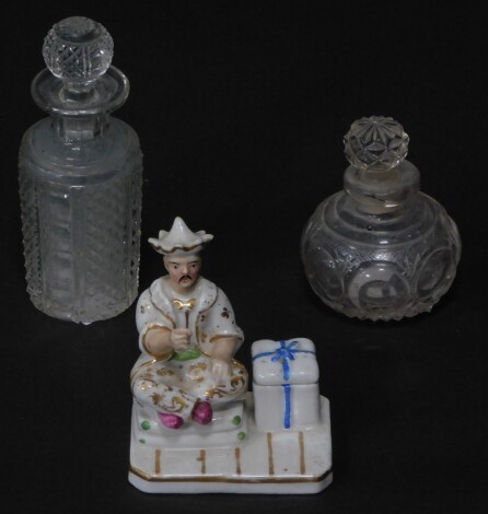 A Continental late 19thC period porcelain figural inkwell, manner of Jacob Petit, modelled with a seated Chinese figure, raised on a canted rectangular base, 8.5cm wide, together with two Victorian cut glass scent bottles and stoppers. (3)