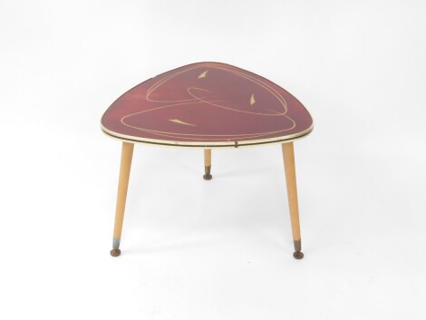 A vintage 1950's occasional table, possibly by Ilse Mobel, Denmark, the curved triangular top set with fibre glass, with stylized gilt decoration against a red ground, the sides with brass effect plastic binding, raised on three turned oak legs, metal cap