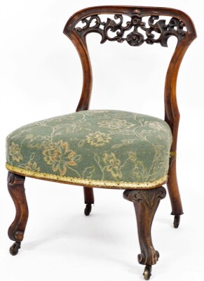 A Victorian carved mahogany nursing chair, the wooden back with fleur de lys and floral carving, on a green rough material seat, with shell legs and castors, 69cm high, 47cm wide, 44cm deep.