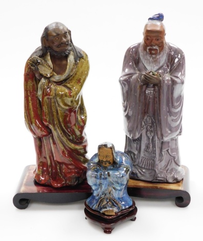 Three Chinese glazed pottery "mudmen" Immortals, each with glazed colouring, including a sage with purple robes, 34cm high, a bearded figure holding a shoe in red and green robes, 32cm high, and another sage robed in blue, hardwood bases, 13cm high. (3)