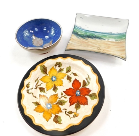 A Highland stoneware pottery dish, painted with a coastal scene, 18cm wide, a Gouda pottery plate decorated in the Anita pattern, and an Island of Syke pottery bowl decorated with a leaf against a blue ground. (3)