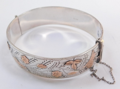 A silver and rose gold plated bangle, engraved and embossed with a bird, butterfly, flowers and leaves, on a snap clasp, with safety chain as fitted, 1.7oz. - 2