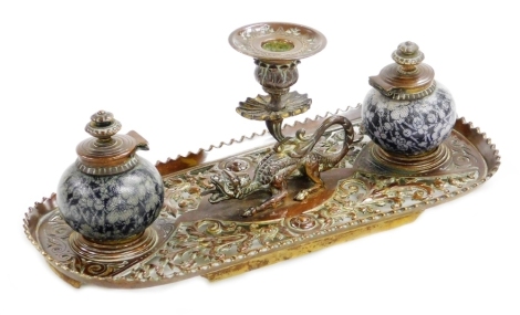 A 19thC continental brass ink stand, with central dragon taperstick and two stoneware inkwells each with textured floral decoration, 32cm wide, 13cm wide, 14cm deep. (AF)