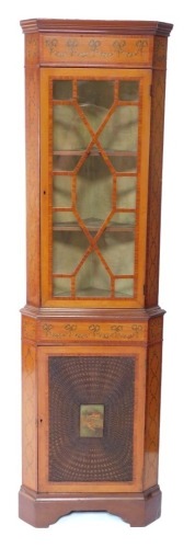 An Edwardian Sheraton Revival satinwood and painted corner display cabinet, painted with floral swags and bows, and interlocking trails of hare bells, the outswept pediment over a glazed door, enclosing two shelves, above a cane paneled door, set with a p