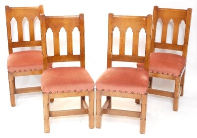 A set of four late 19thC Liberty & Co oak dining chairs, each with a Gothic shaped back, pink upholstered padded seat, on chamferred legs with H stretcher, bone labelled to underside.