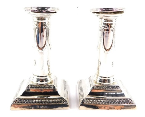 A pair of Edward VII Adam style loaded silver candlesticks, embossed with bows and swags, raised on a stepped square base, London 1905, 24.4oz all in.