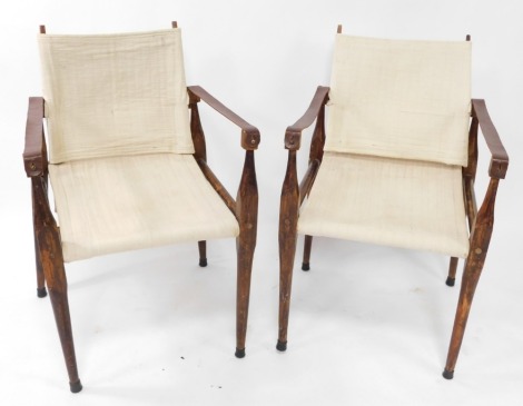 A pair of Anglo Indian stained wood and leather campaign open armchairs, each with a cream upholstered back and seat, on turned tapering legs.