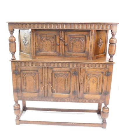 A Jacobean style oak court cupboard, the upper section with double cupboards, beneath a carved canopy cornice held on heavy supports raised above triple cupboards each with heavily carved doors, on heavily carved legs on block feet joined by horizontal bl