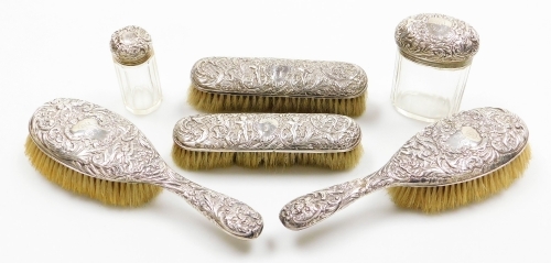 A Victorian silver backed dressing table set, each with heavy foliate scroll detailing with cherubs, bearing the initials MR, comprising two hand brushes, London 1901, two smaller brushes London 1902 and two silver topped glass dressing table jars, London