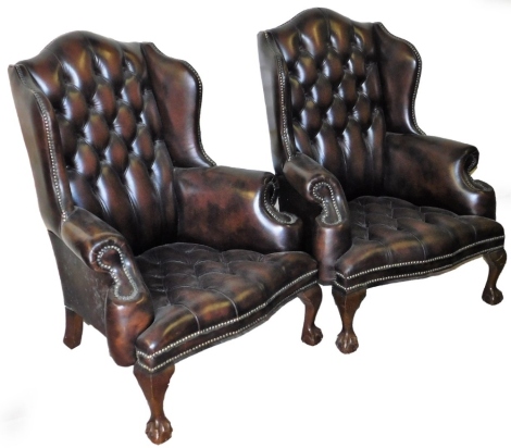 A pair of Chesterfield style wing back armchairs, each upholstered in a brown studded leather, with ball and claw feet, 102cm high, 75cm wide, 56cm deep.