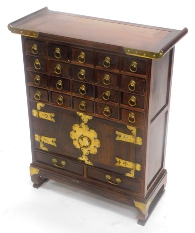 A Chinese hardwood and brass side cabinet, with an arrangement of small drawers and two doors, each with strap hinges on bracket feet, 61cm wide.