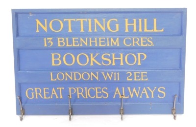 A painted wood and metal wall mounted coat rack, the rectangular panel board painted with "Nottinghill Book Shop, 13 Blenheim Crescent, London, W11 2EE, Great Prices Also", the four coat hooks of scrolling form, 65cm high, 101cm wide.