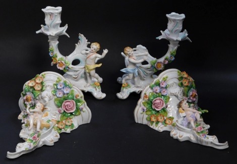 A pair of Sitzendorf 20thC porcelain wall shelves, of rococo form, encrusted with flowers and surmounted with cherubs, 16.5cm high, together with a pair of rococo style figural candlesticks, with putti and encrusted with flowers, 18cm high. (4)