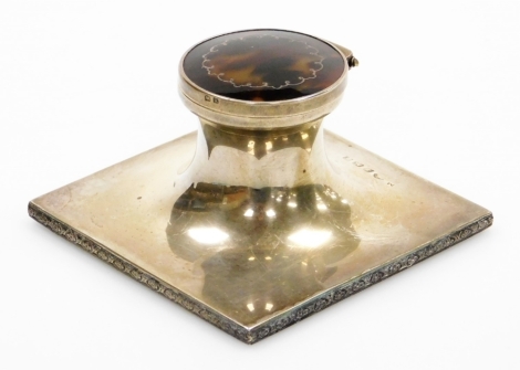 A George V silver and tortoiseshell inkwell, the circular inkwell top with tortoiseshell inlay and applied silver detailing, with a glass inset inkwell on a square base with foliate design border, maker Adie Bros Ltd, Birmingham 1925, 5.5cm high, on a wei