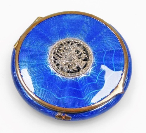 An early 20thC blue guilloche enamel decorated powder compact, with a silver coloured metal flower basket inset to the lid, with an outer plated border, the hinged lid enclosing a mirror back and a gilt metal rotating powder compartment, 5.5cm diameter. (