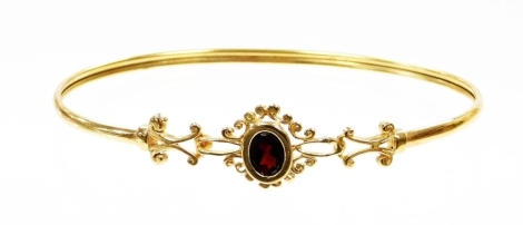 A 9ct gold hinged bangle, with central oval cut garnet in floral cluster setting with scroll design shoulders, the clip clasp opening passed the central floral cluster, 6.5cm diameter, 4.4g all in.