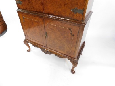 A mid century burr walnut cocktail cabinet, the foliate carved top over a pair of doors, opening to reveal a mirror back, glass shelves and fitted interior, above a further door enclosing a drawer and shelves, on cabriole legs, 159cm high, 85cm wide, 41cm - 5