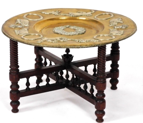 An Eastern brass tray top table, the brass charger embossed with flowers, elephants, lions, etc., on a mahogany folding stand, on turned legs, the charger 60cm diameter.